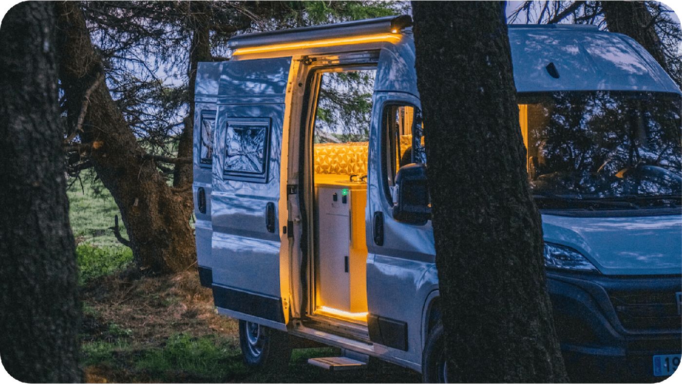 Dos furgonetas camper aparcadas entre árboles al anochecer, con la puerta lateral abierta y la luz interior encendida.