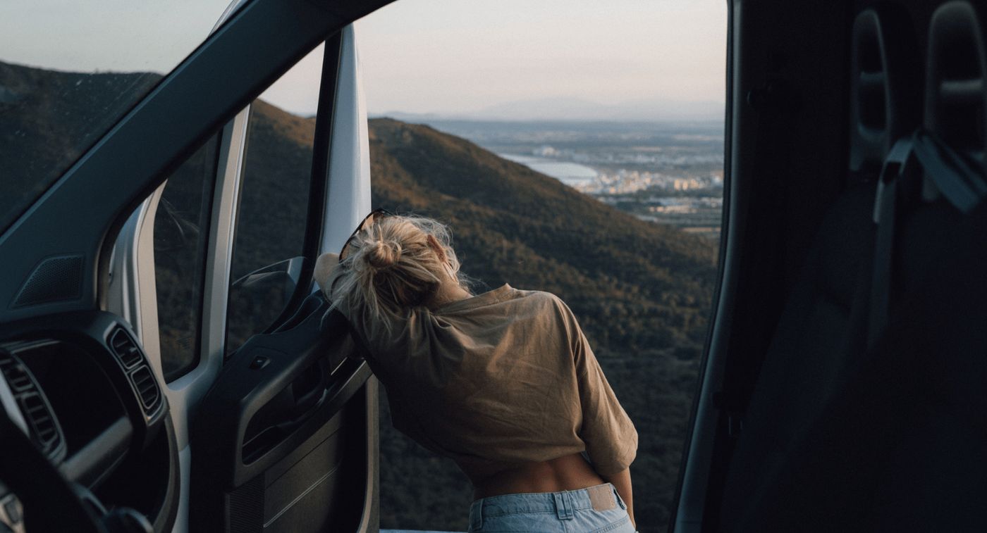 Chica de espaldas apoyada en la puerta de su camper disfrutando de las vistas al aire libre.