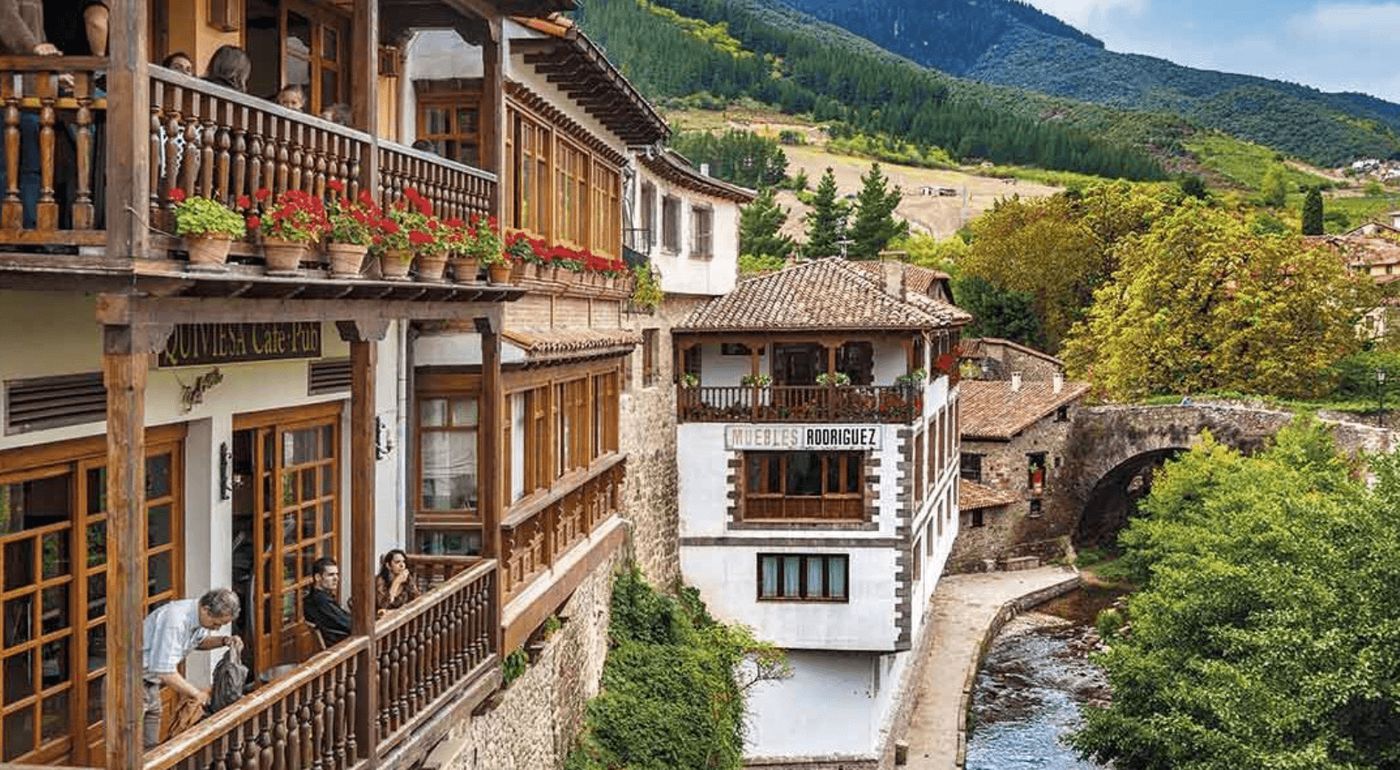 Coloridas calles de un pintoresco pueblo de Liébana, con un río y puente al fondo, ideales para explorar durante una ruta en furgoneta camper por los Picos de Europa.